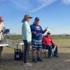 Barb Semeraro being welcomed into the flying competition by Henry Toews with a bottle of wine, a jet cat kit, and an award. Arnold is distracted and Hodes is exhausted.